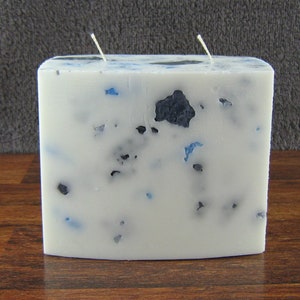 May include: A square, white candle with two wicks. The candle is speckled with blue and black decorative elements. The candle sits on a wooden surface.