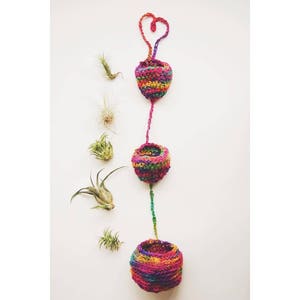 May include: A set of three colorful crocheted plant hangers with a heart-shaped top. Each hanger is filled with a small air plant.
