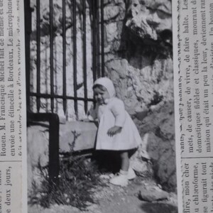 Puede incluir: Una fotograf&iacute;a en blanco y negro de una ni&ntilde;a peque&ntilde;a que lleva un vestido blanco y un gorro blanco. Est&aacute; de pie frente a una puerta de metal y mirando hacia la izquierda. La foto est&aacute; ligeramente descolorida y tiene un aspecto vintage.