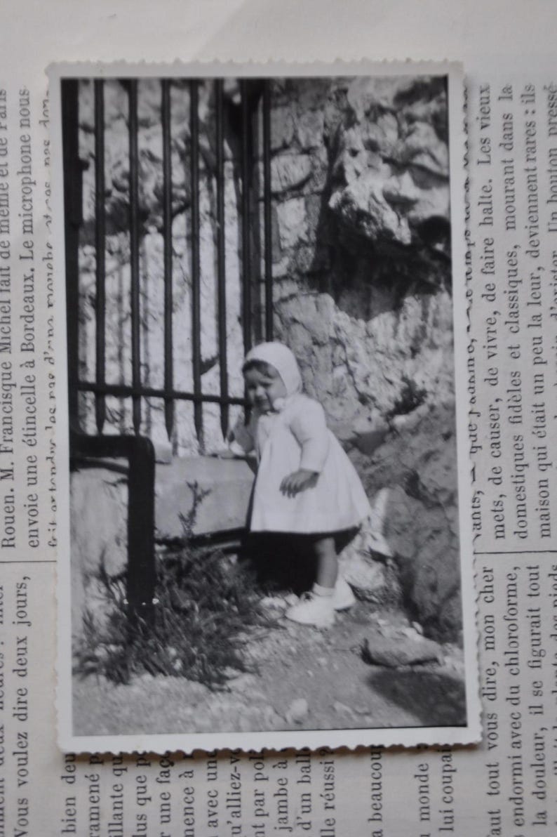 Puede incluir: Una fotograf&iacute;a en blanco y negro de una ni&ntilde;a peque&ntilde;a que lleva un vestido blanco y un gorro blanco. Est&aacute; de pie frente a una puerta de metal y mirando hacia la izquierda. La foto est&aacute; ligeramente descolorida y tiene un aspecto vintage.