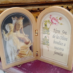 Pode incluir: Uma moldura de madeira arqueada com dois painéis. Um painel apresenta uma estátua de uma mulher e uma criança. O outro painel tem texto e um desenho floral. O texto diz "Ntra. Sra. de la Oliva bendice a nuestra familia."