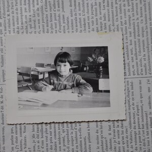 Puede incluir: Una fotograf&iacute;a en blanco y negro de una ni&ntilde;a sentada en un escritorio en un aula. Lleva una camisa de color claro y tiene el pelo recogido hacia atr&aacute;s. Est&aacute; mirando un libro o un cuaderno.