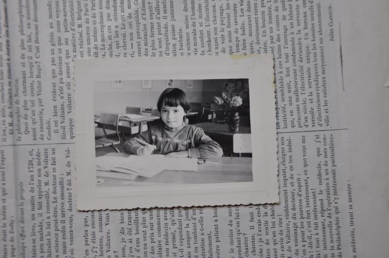 Puede incluir: Una fotograf&iacute;a en blanco y negro de una ni&ntilde;a sentada en un escritorio en un aula. Lleva una camisa de color claro y tiene el pelo recogido hacia atr&aacute;s. Est&aacute; mirando un libro o un cuaderno.