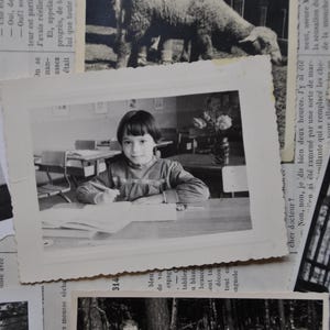 Puede incluir: Una fotograf&iacute;a en blanco y negro de una ni&ntilde;a sentada en un escritorio en un aula. Lleva una camisa de color claro y tiene el pelo recogido hacia atr&aacute;s. Est&aacute; mirando un libro delante de ella.