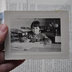 Puede incluir: Una ni&ntilde;a joven con cabello oscuro se sienta en una mesa en un aula. Lleva un su&eacute;ter de color claro y mira un libro. La mesa est&aacute; cubierta de papeles y libros.