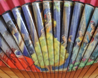 May include: A red and wooden folding fan with a painted design of women in colorful dresses. The fan has a wooden handle and is decorated with yellow and white painted flowers.