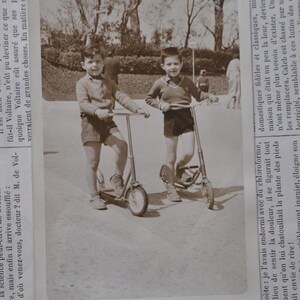 Puede incluir: Dos ni&ntilde;os peque&ntilde;os con pantalones cortos y camisetas montan en patinete en un parque. Los ni&ntilde;os sonr&iacute;en y parecen divertirse.