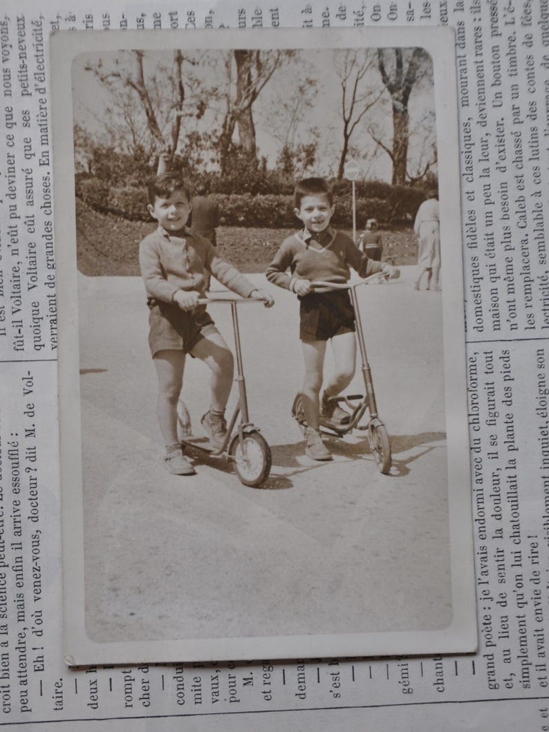 Puede incluir: Dos ni&ntilde;os peque&ntilde;os con pantalones cortos y camisetas montan en patinete en un parque. Los ni&ntilde;os sonr&iacute;en y parecen divertirse.