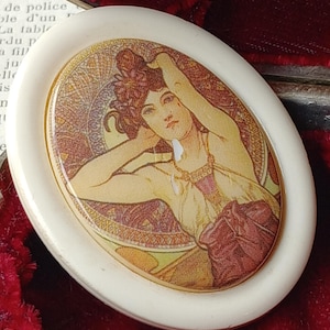 May include: A white oval-shaped trinket box with a raised rim. The lid features a colorful Art Nouveau style image of a woman with flowing hair and a pearl necklace. The image is set against a background of swirling patterns in shades of gold, brown, and red.