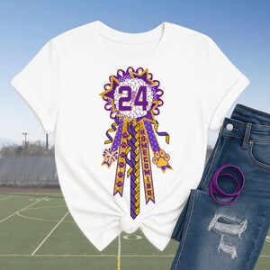 May include: White t-shirt featuring a purple and gold homecoming ribbon with the number 24. The ribbon has a starburst design, paw prints, and the word "HOMECOMING". The shirt is paired with blue jeans and a purple hair tie.