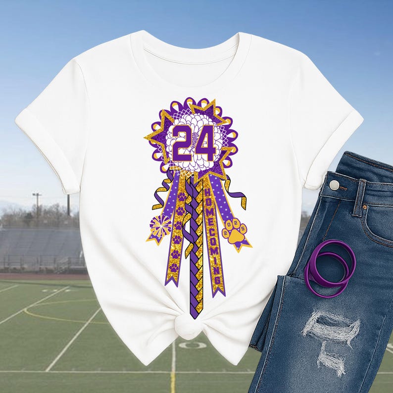 May include: White t-shirt featuring a purple and gold homecoming ribbon with the number 24. The ribbon has a starburst design, paw prints, and the word "HOMECOMING". The shirt is paired with blue jeans and a purple hair tie.