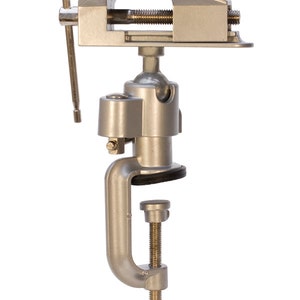 May include: A silver-colored bench vise with a clamp base and adjustable jaws. The vise has a swivel base and a handle for tightening. The jaws are lined with black rubber pads. This tool is designed for holding objects securely during various tasks.