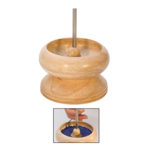 May include: A wooden bead spinner with a stainless steel needle. The spinner has a double-bowl design, with a wider top bowl and a smaller bottom bowl. The wood has a light, natural finish. A smaller inset image shows the spinner in use with blue beads.