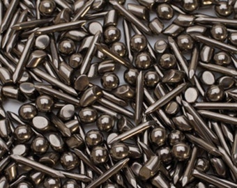 May include: An overhead shot displays a dense collection of small, metallic objects. The assortment includes rounded spheres, cylindrical pieces, and various shapes with pointed ends. The objects have a dark, gunmetal finish, creating a uniform, industrial aesthetic.