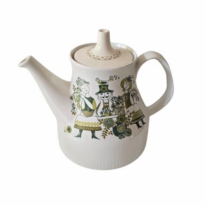 May include: White ceramic teapot with a green and brown floral design featuring three figures. The teapot has a lid and a handle.
