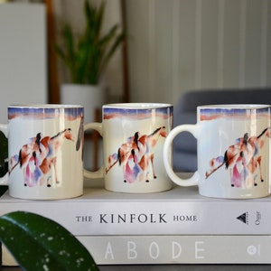 May include: Three white ceramic mugs with a colorful design of a horse and people. The mugs are on top of two books with the text "The Kinfolk Home Abode".