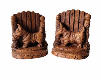 May include: A pair of wooden bookends featuring a Scottish Terrier dog standing in front of a wooden fence. The bookends are brown and have a rustic look.