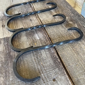 May include: Four black metal S-hooks with a twisted center design. The hooks are arranged on a weathered wooden surface, showcasing their curved shape and dark color. These hooks are ideal for hanging items.