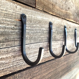 May include: A row of four black, hand-forged metal hooks mounted on a weathered wooden surface. Each hook is a simple, curved design, attached with a single screw. The wood has a rustic, aged appearance.