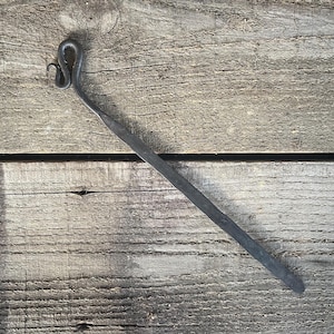 May include: Hand-forged, dark gray metal fire poker with a curved handle and a long, tapered shaft. The poker is lying on a weathered, wooden surface, showcasing its rustic, handcrafted appearance.