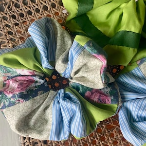 Puede incluir: Una gran flor de tela multicolor con un centro marrón. La flor está hecha de diferentes telas estampadas, incluyendo flores, rayas y colores sólidos. La flor está sentada sobre una superficie de mimbre tejido.