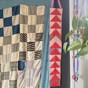 Könnte beinhalten: Eine Patchwork-Decke mit verschiedenen Stoffen und Mustern, darunter Gingham, Punkte und Streifen. Die Decke hängt an einer schwarzen Wand mit einem rosa und roten Stoffstreifen daneben.