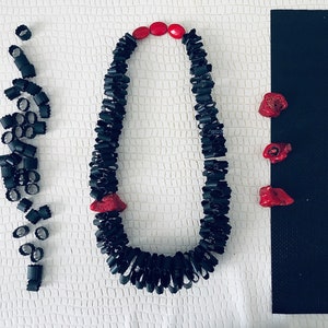 May include: A necklace composed of black cylindrical beads and three bright red organic-shaped accents, arranged in a loop. To the left are loose black cylindrical beads, and to the right are three separate red organic-shaped pieces.