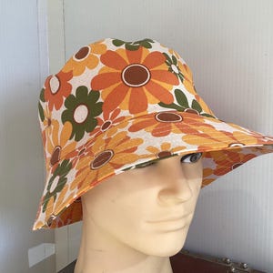 May include: A bucket hat with a floral pattern in a retro style. The hat displays large orange, yellow, and green flowers on a cream background. The brim is wide. The hat is displayed on a mannequin head.