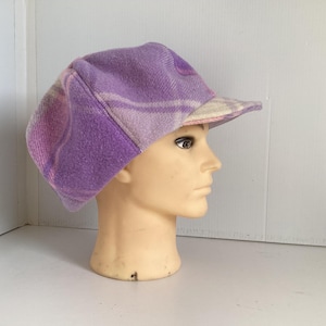 May include: A newsboy cap featuring a soft, felted texture in shades of lavender and light purple with a subtle plaid pattern. The cap has a rounded, multi-paneled crown and a short, stiffened brim that extends from the front of the hat.