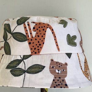 May include: A white bucket hat featuring a repeating pattern of cheetahs, leaves, and giraffe illustrations in brown, black, and green. The hat has a wide brim and is made of a soft fabric.