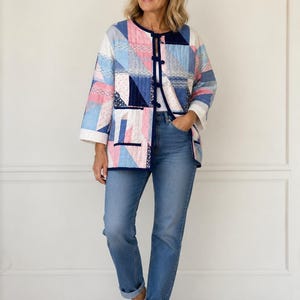 Könnte beinhalten: Eine Patchwork-Jacke in Blau-, Rosa- und Weißtönen mit geometrischem Design. Die Jacke hat eine marineblaue Paspelierung und einen Knopfverschluss. Das Model trägt eine blaue Jeans und weiße Sneaker.
