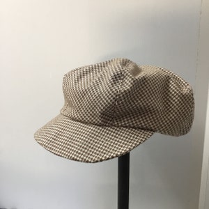 May include: A brown and white houndstooth pattern newsboy cap. The cap features a rounded crown and a short, curved brim. The fabric is a tightly woven material. The cap is displayed on a black stand against a white background.
