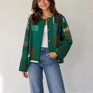 May include: A green patchwork jacket with gold button fastenings. The jacket incorporates various fabric patterns and textures. The person is wearing blue jeans and a white top.