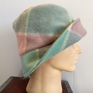 May include: A bucket hat featuring a soft, felted texture with a large-scale plaid pattern in muted shades of sage green, dusty pink, and light grey. The hat has a rounded crown and a downward-sloping brim that is slightly turned up at the front.