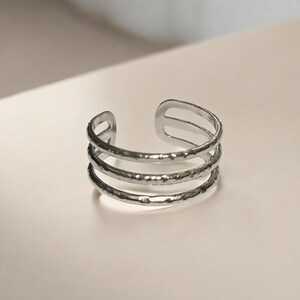 May include: A silver ring with three textured bands. The open-ended design features three bands with a hammered or irregular surface. The ring is likely made of metal and has a simple, modern aesthetic. The bands are approximately 0.5 cm wide.