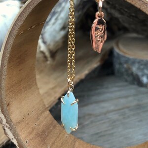 May include: A gold chain necklace with a light blue crystal pendant. The pendant is faceted and has a gold setting. The necklace is hanging from a wooden surface.