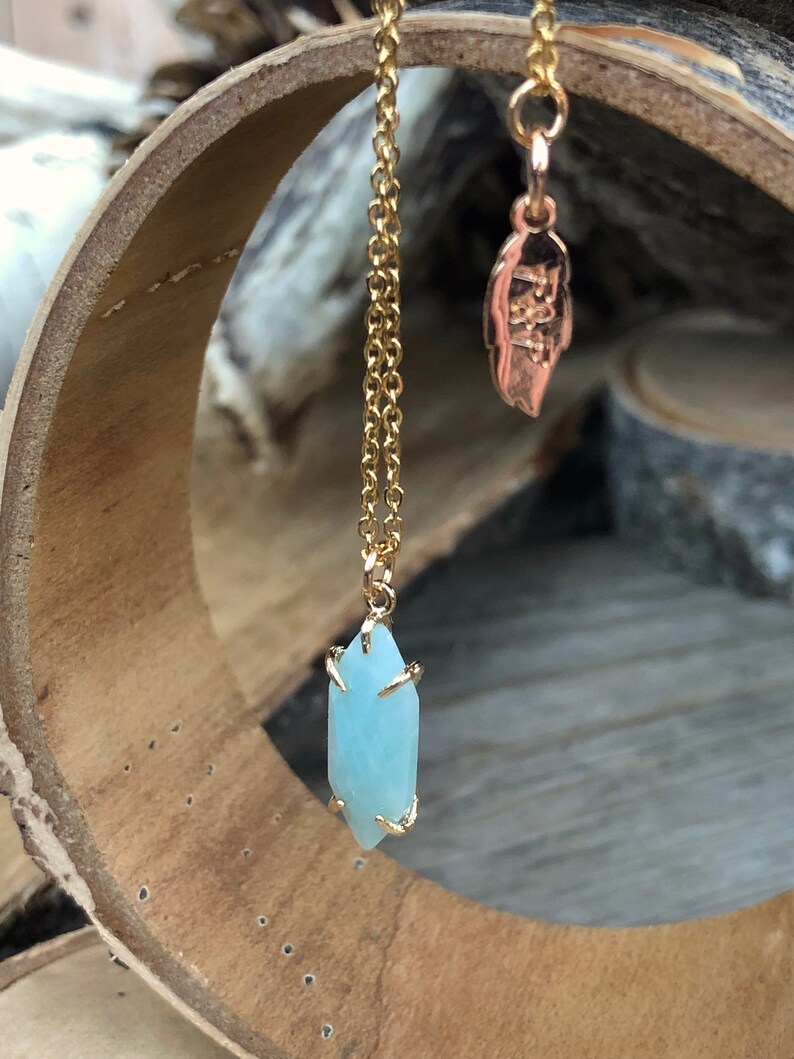 May include: A gold chain necklace with a light blue crystal pendant. The pendant is faceted and has a gold setting. The necklace is hanging from a wooden surface.