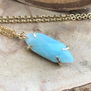 May include: A gold chain necklace with a light blue gemstone pendant. The pendant is a faceted, elongated shape with gold accents.