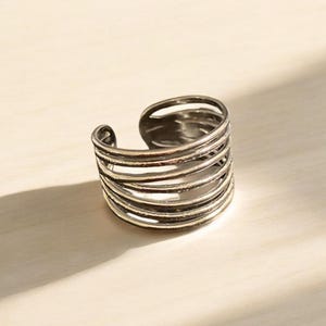 Op de afbeelding: Zilverkleurige verstelbare ring met een meerlaags ontwerp. De ring heeft een reeks horizontale banden, waardoor een textuur en open uitstraling ontstaat. De ring staat op een lichtgekleurd oppervlak, met schaduwen die diepte toevoegen.