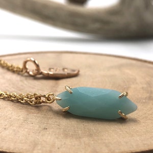 May include: A gold chain necklace with a light blue gemstone pendant. The pendant is a faceted, elongated oval shape with gold accents.
