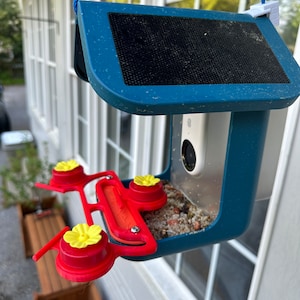 May include: A blue bird feeder with a solar panel on top and a clear plastic feeder section. The feeder has three red perches with yellow flower-shaped tops. The feeder is hanging from a blue rope.