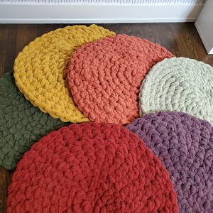 Pu&ograve; includere: Un set di sei cuscini rotondi in crochet per il pavimento in varie tonalit&agrave; di rosso, arancione, giallo, verde e viola. I cuscini sono disposti a semicerchio su un pavimento in legno.