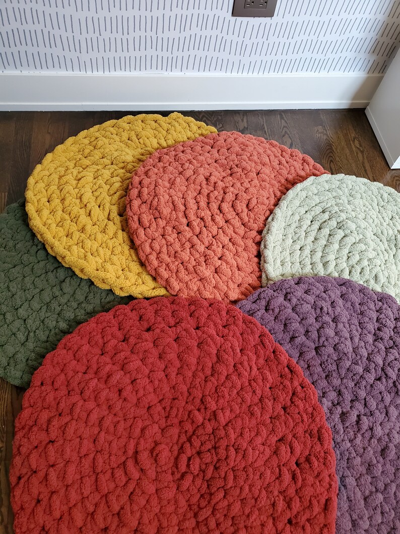 Pu&ograve; includere: Un set di sei cuscini rotondi in crochet per il pavimento in varie tonalit&agrave; di rosso, arancione, giallo, verde e viola. I cuscini sono disposti a semicerchio su un pavimento in legno.