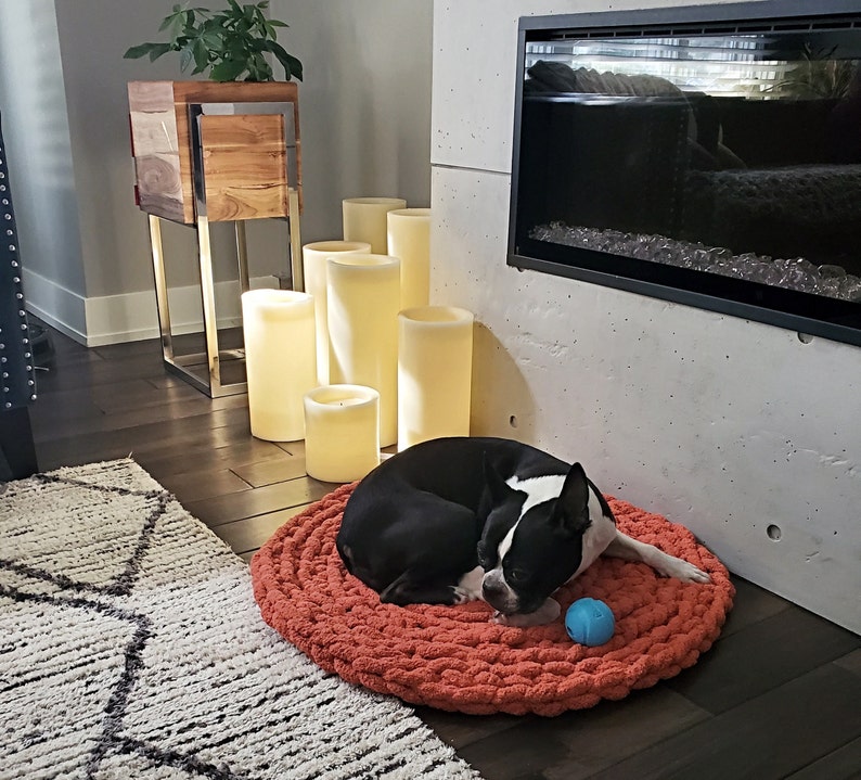 Pu&ograve; includere: Un Boston Terrier bianco e nero sta dormendo su un letto per cani in crochet color arancio bruciato con una palla giocattolo blu. Il letto per cani &egrave; su un pavimento in legno duro davanti a un camino con candele.