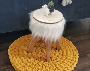May include: A white furry stool with wooden legs sits on a round, mustard yellow crocheted rug. A small succulent plant in a black pot sits on top of the stool.