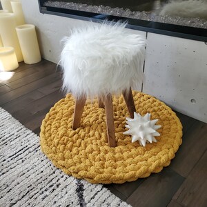 Pu&ograve; includere: Uno sgabello bianco in pelliccia sintetica con gambe in legno si trova su un tappeto rotondo di maglia gialla. Un oggetto decorativo bianco con punte si trova sul tappeto.