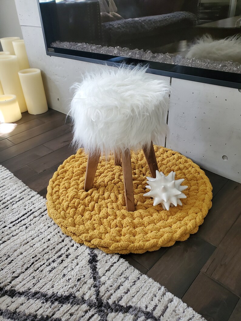 Pu&ograve; includere: Uno sgabello bianco in pelliccia sintetica con gambe in legno si trova su un tappeto rotondo di maglia gialla. Un oggetto decorativo bianco con punte si trova sul tappeto.