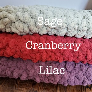 Pu&ograve; includere: Tre cuscini da divano in maglia di colore salvia, mirtillo e lilla. I cuscini sono realizzati in filato spesso e hanno una consistenza soffice.