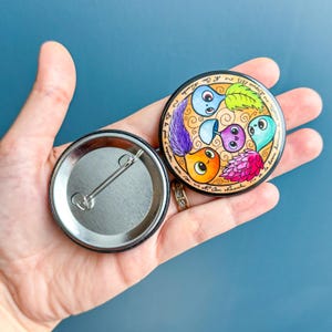 Puede incluir: Un botón redondo con una ilustración colorida de cinco personajes de dibujos animados con un mensaje escrito a mano alrededor del borde. El botón se sostiene en una mano. La parte posterior del botón es plateada con un pasador de seguridad.