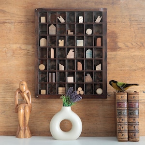 May include: A vintage printer's tray with various wooden shapes. A white, ring-shaped vase with lavender. A carved wooden figure and a bird ornament are placed near two antique books. The scene is set against a wooden backdrop.